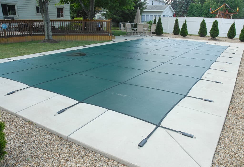 A green DLI pool safety cover in place covering a rectangular pool in a backyard.